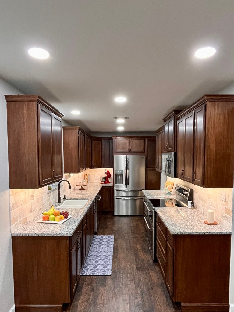 Kitchen with dark stained wood cabinets, light cream granite countertops. It is a galley style kitchen with the fridge at the back of the photo.