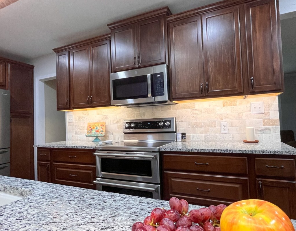 Kitchen with dark stained wood cabinets, light cream granite countertops. The right side of the kitchen. In the foreground there is fresh grapes and an apple. It also shows a stainless steel range with a double oven and a stainless steel microwave above it. There is an opening on the far left of the photo that takes you to more of the house.