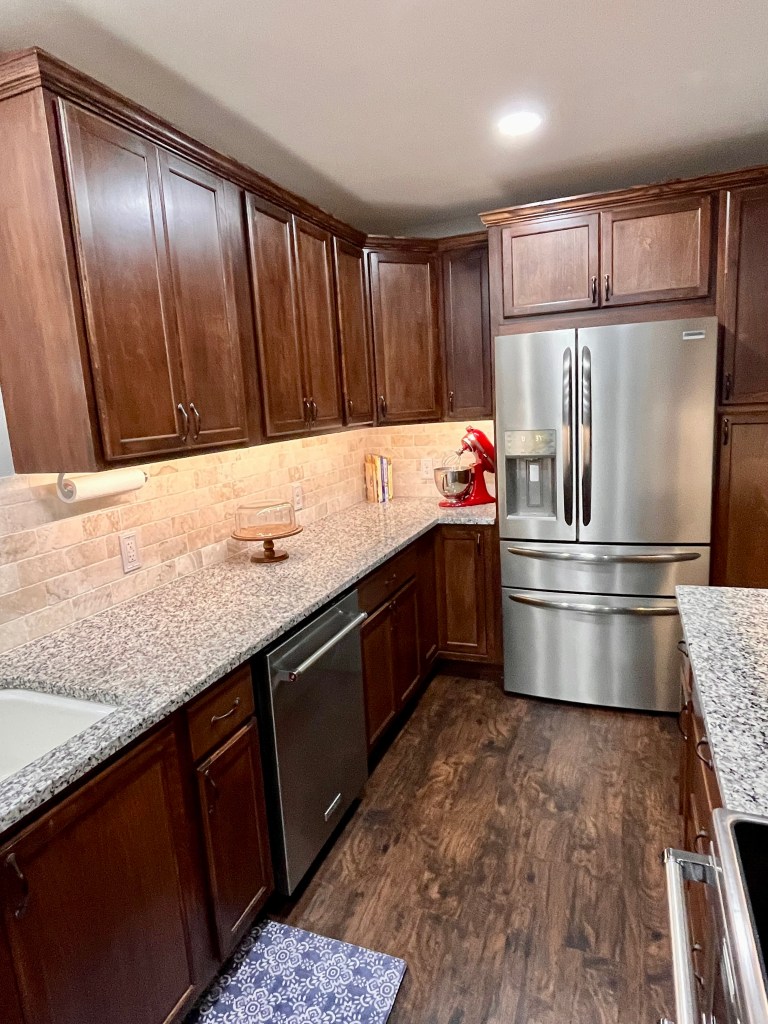Kitchen with dark stained wood cabinets, light cream granite countertops. The left side of the kitchen and the fridge at the back of the kitchen. There is a cake stand and red KitchenAid mixer on the counter. There is a stainless steel dishwasher to the right of the sink.