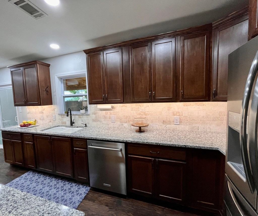 Kitchen with dark stained wood cabinets, light cream granite countertops. The left side of the kitchen. There is a window centered over the sink, a stainless steel dishwasher to the right of the sink, and there is a cake stand on the counter just to the right of the dishwasher.