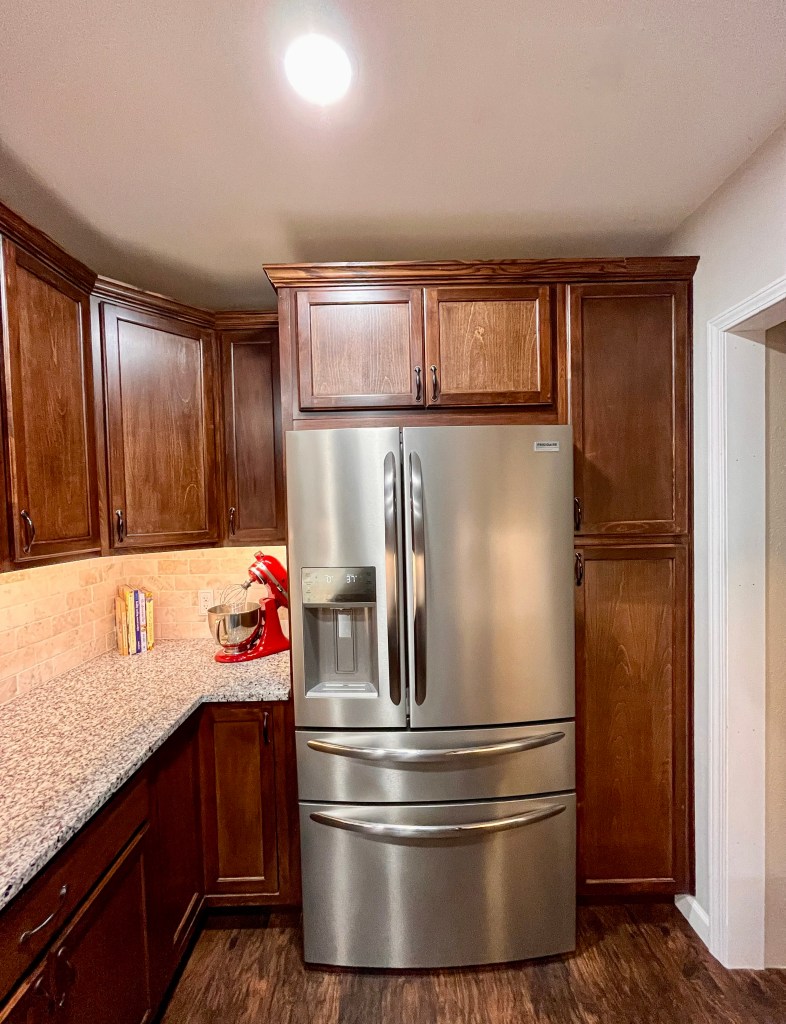 Kitchen with dark stained wood cabinets, light cream granite countertops. Close-up of the back of the kitchen. There is countertops on the left with a KitchenAid mixer on top of it. The countertops abut up to a stainless steel fridge. It has double doors up top, a small drawer beneath the doors, and a big drawer at the bottom.