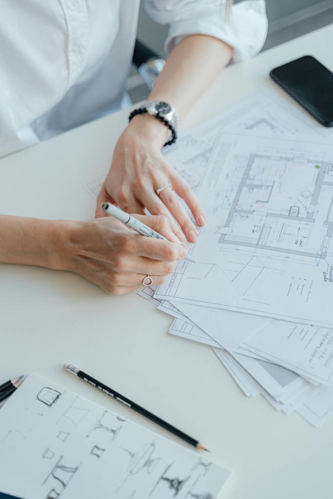 An interior designer drafting a floor plan that is on top of other floor plans. There is small out of focus sketches, a pencil and pen, all on a white table. There is a phone next to a woman's left arm that has a watch and black bracelet on it. She is holding a pen and wearing a white button up shirt. 